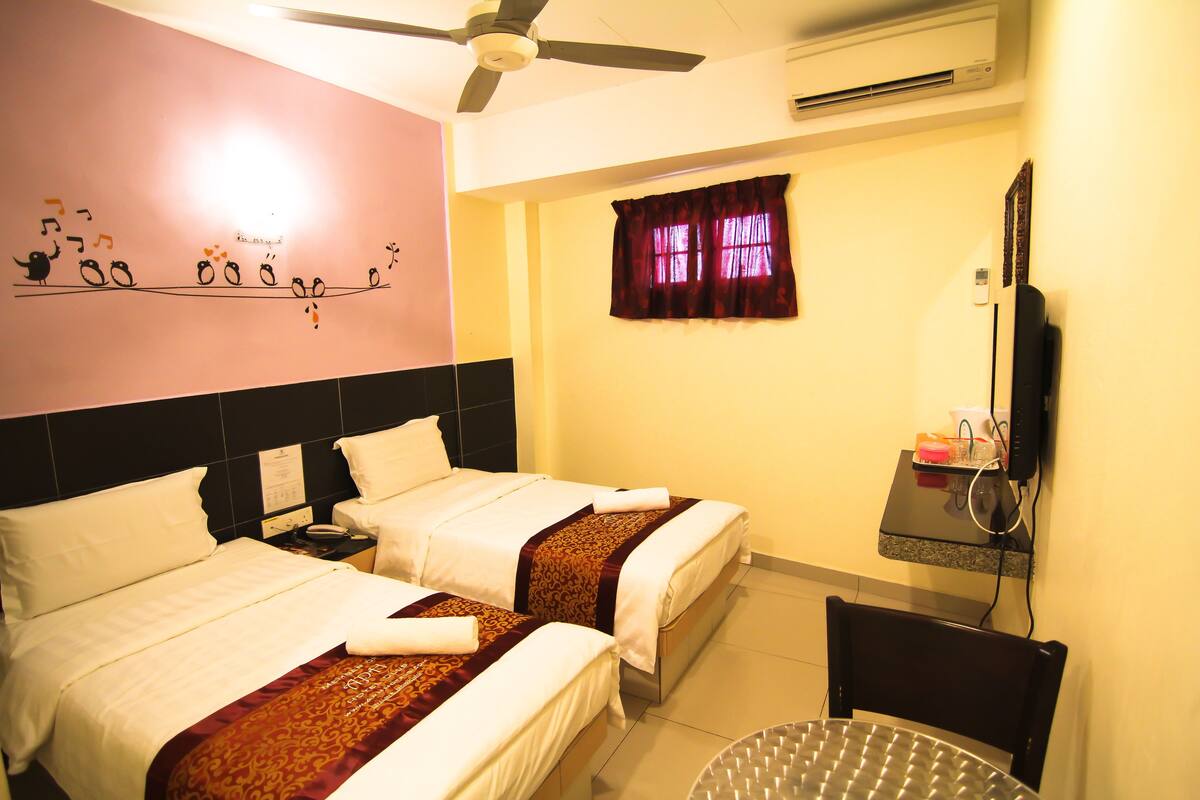 A hotel room with two beds, a ceiling fan, an air conditioner, a TV mounted on the wall, and a small table with a chair.