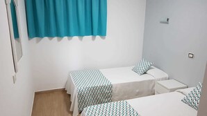 Una habitación pequeña y limpia con dos camas individuales, una ventana con cortina color turquesa y una pared blanca.
