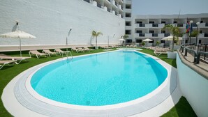 Una piscina con sillones de descanso y sombrillas, rodeada por un edificio blanco con balcones.