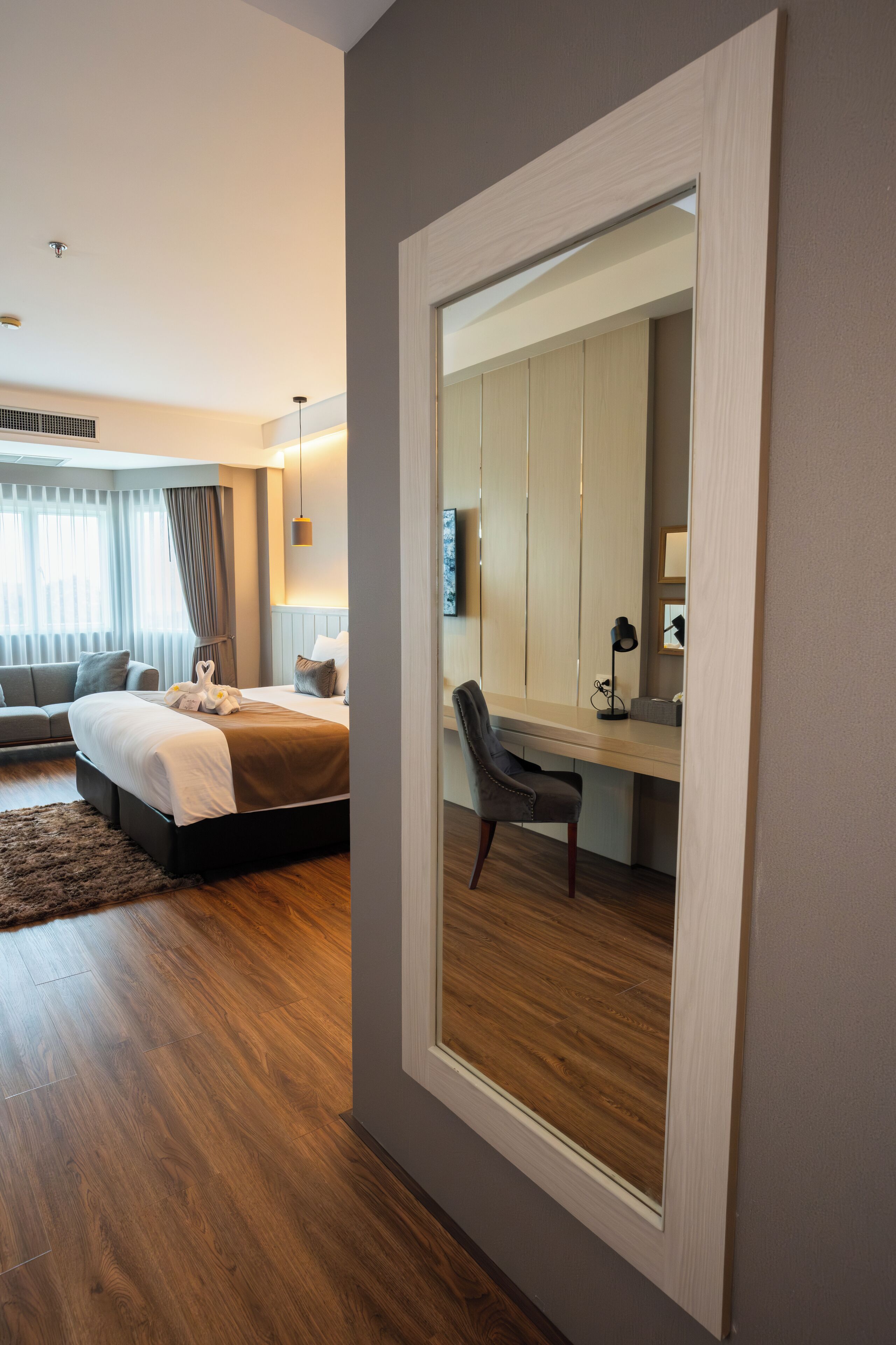 A modern hotel room with a large bed, a desk with a chair, a large mirror, and a view of the city through the window.