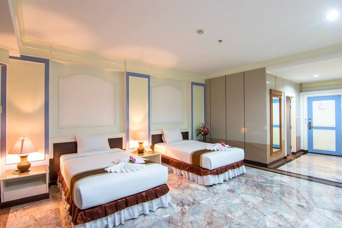 Una habitación de hotel con dos camas, una mesita de noche con una lámpara, un espejo grande y una puerta que da a otra habitación.