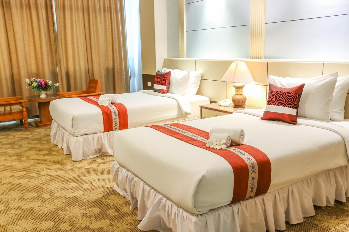 A hotel room with two beds, a desk, a chair, a lamp, and a vase with flowers.