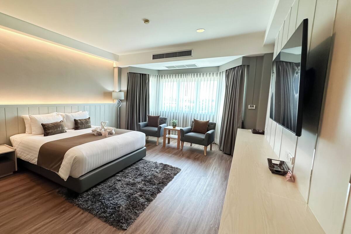 Una habitación de hotel moderna con una cama grande, un escritorio, dos sillones, un televisor de pantalla plana y un ventanal con cortinas.