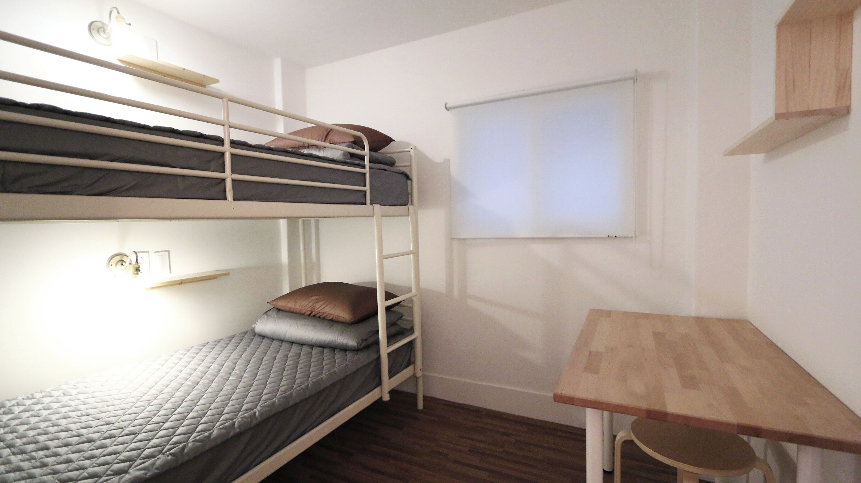 A bunk bed room with a desk and chair, a window, and a wooden shelf.