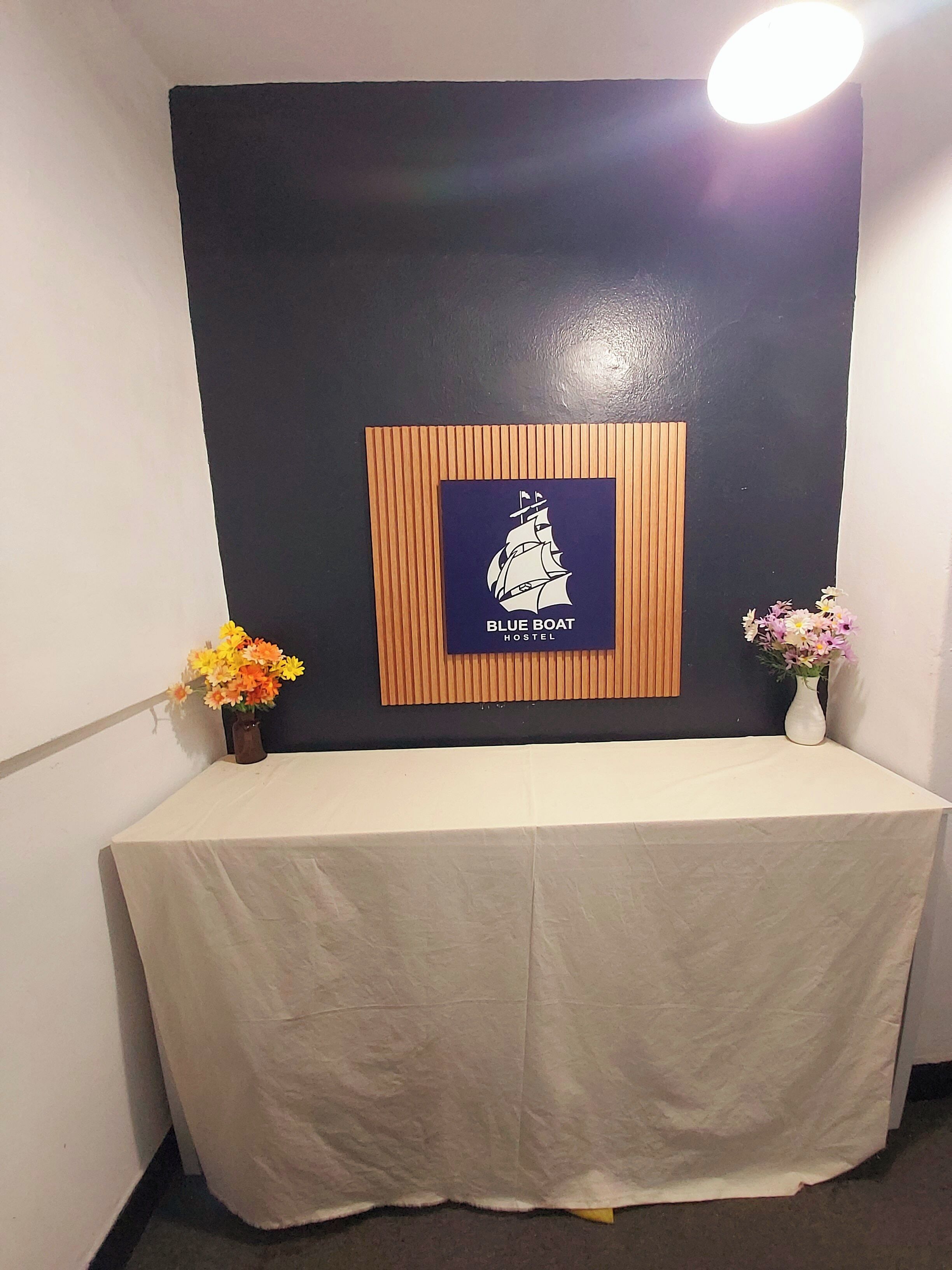 A reception area with a table covered in a white tablecloth, a framed sign reading "BLUE BOAT HOSTEL," and two vases with flowers on either side.