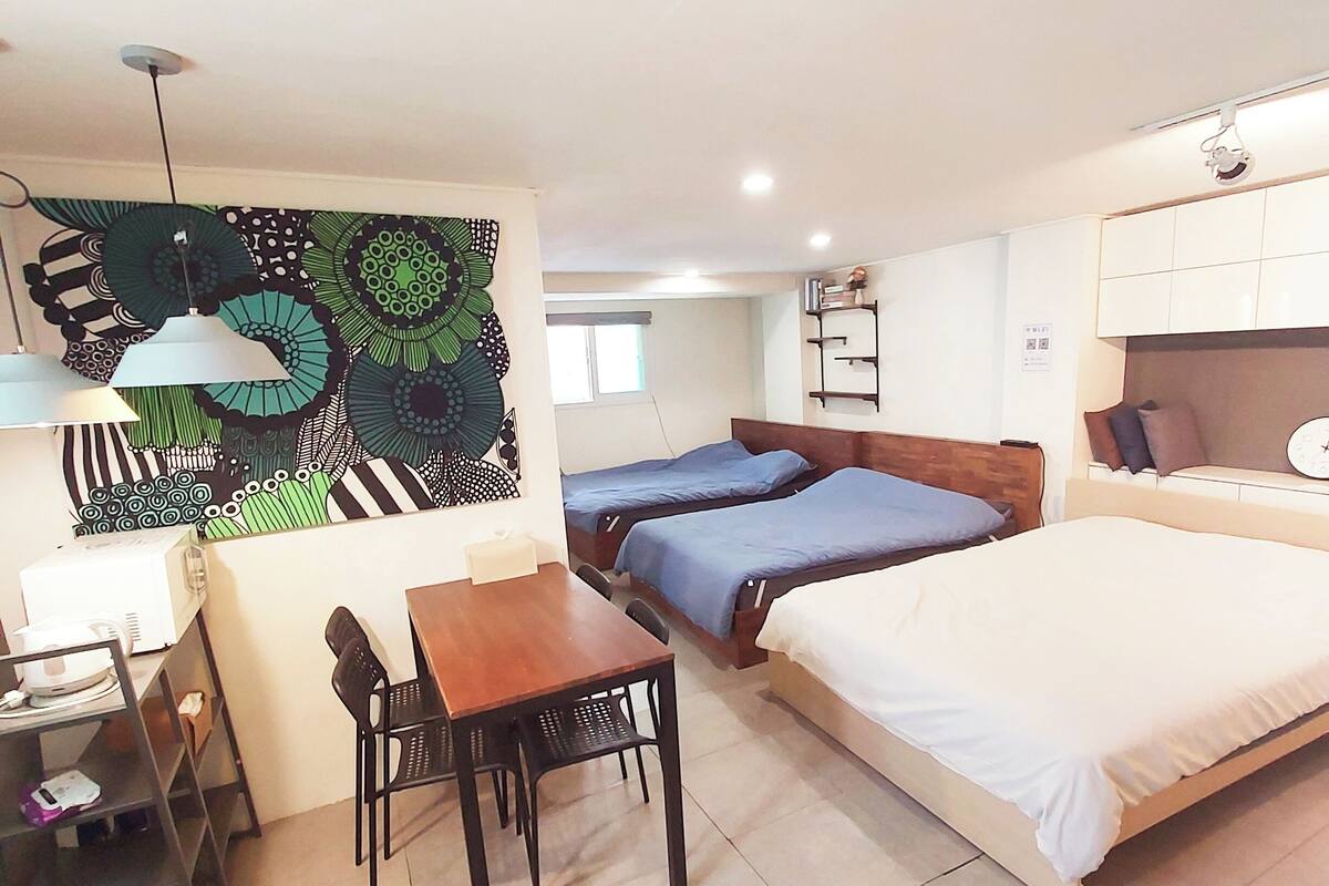 A compact hotel room with a bed, two single beds, a dining table with chairs, a small kitchen area, and a decorative wall piece.