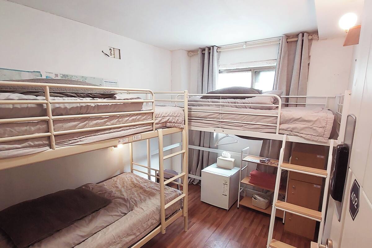 A bunk room with wooden flooring, bunk beds, a small white cabinet, and a window with curtains.