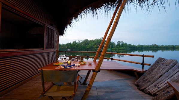 Une terrasse en bois avec une table dressée pour le repas, offrant une vue sur un lac paisible et une végétation luxuriante.