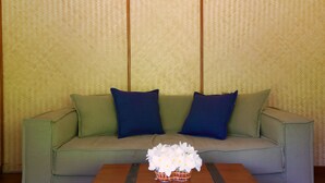 A green sofa with two dark blue pillows, a wooden coffee table with a white flower arrangement, and a patterned wallpaper background. - Yathra Houseboat (Bentota)