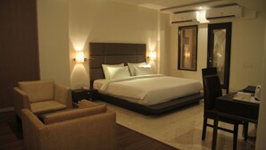 Quarto de hotel com uma cama grande, duas luminárias de cabeceira, uma escrivaninha com cadeira e um sofá.