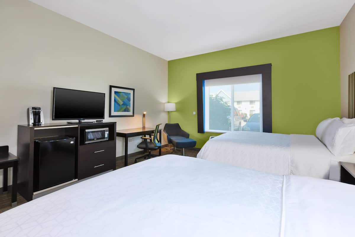 A hotel room with two beds, a desk with a flat-screen TV, a coffee maker, a chair, and a window with a view of buildings.