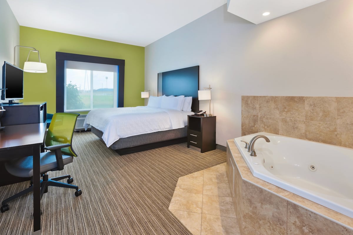 A modern hotel room with a large bed, a desk with a computer, a chair, a large window, a bathroom with a bathtub, and a green accent wall.