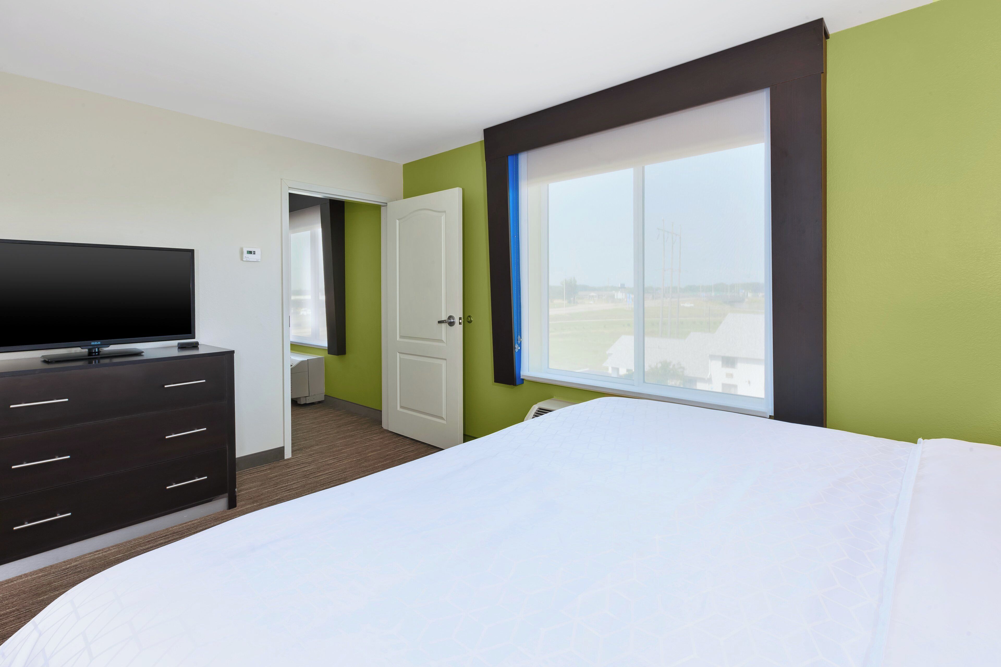 A hotel room with a bed, a television on a dresser, a door, and a window with a view.