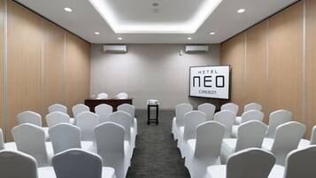 A conference room with white chairs, a wooden desk, and a projector screen displaying "HOTEL NEO CIREBON".