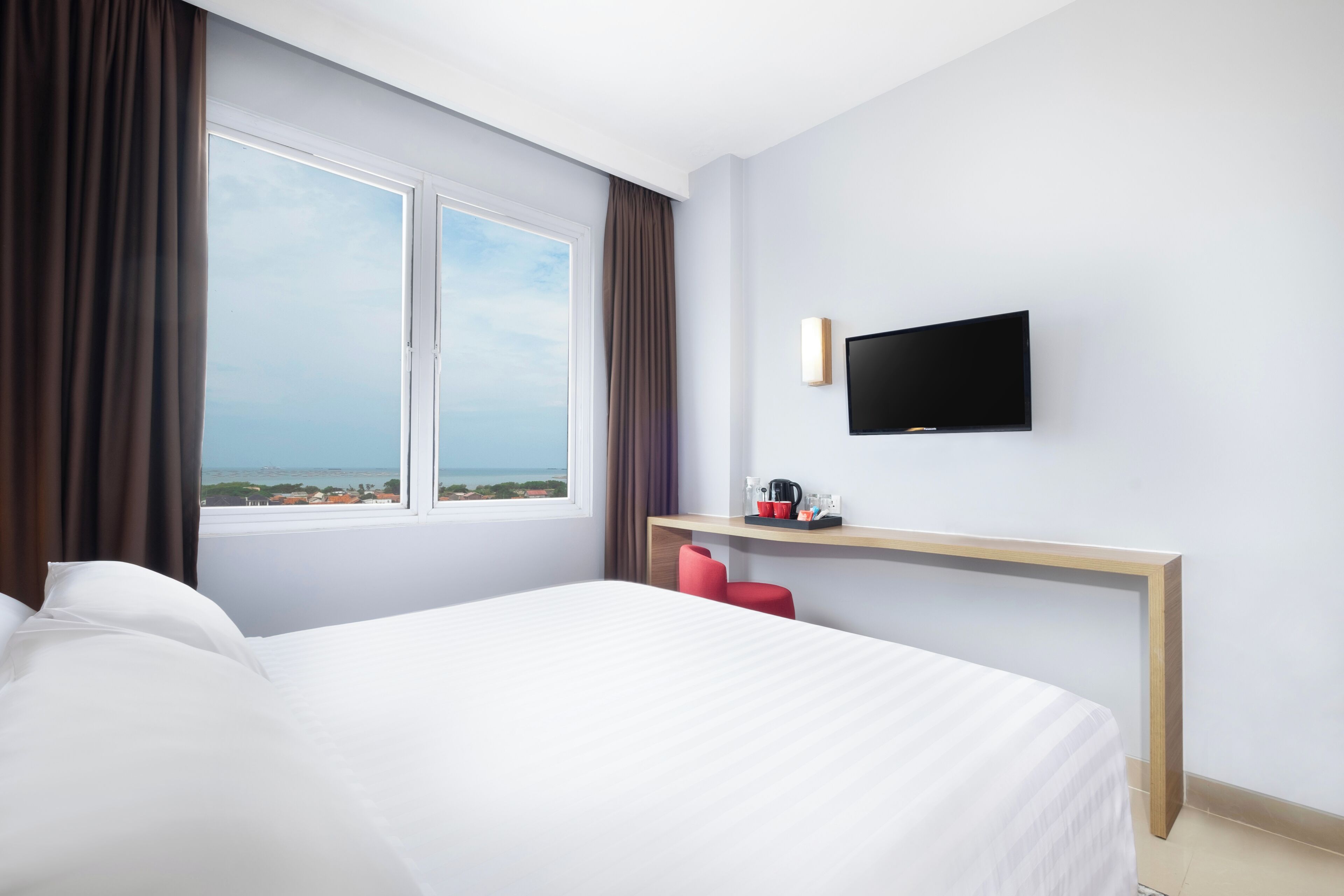 A hotel room with a large bed, a TV mounted on the wall, a desk with a chair, and a window with curtains.