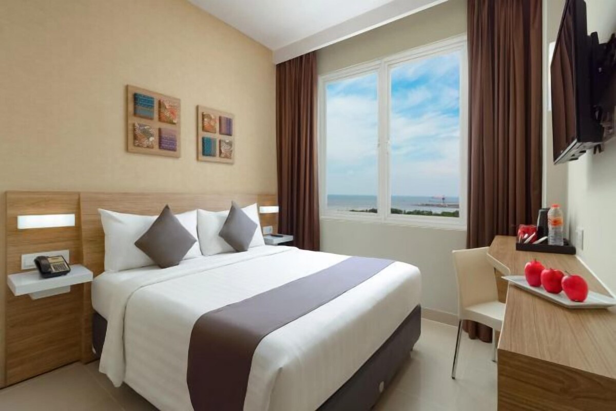 A hotel room with a bed, a desk, a chair, a TV, and a window with a view of the sea.