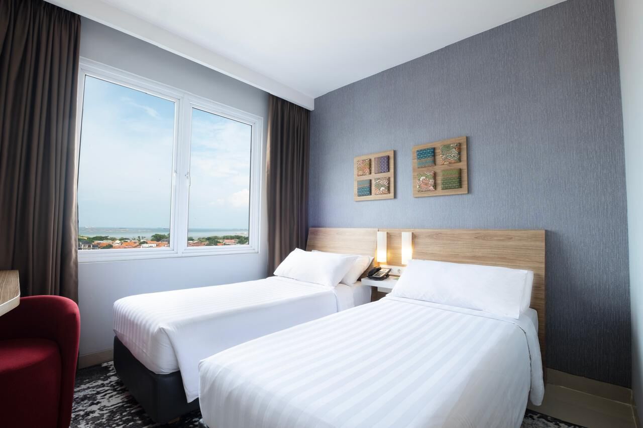 A hotel room with two beds, a desk, a chair, a window with a view, and two wall-mounted artworks.