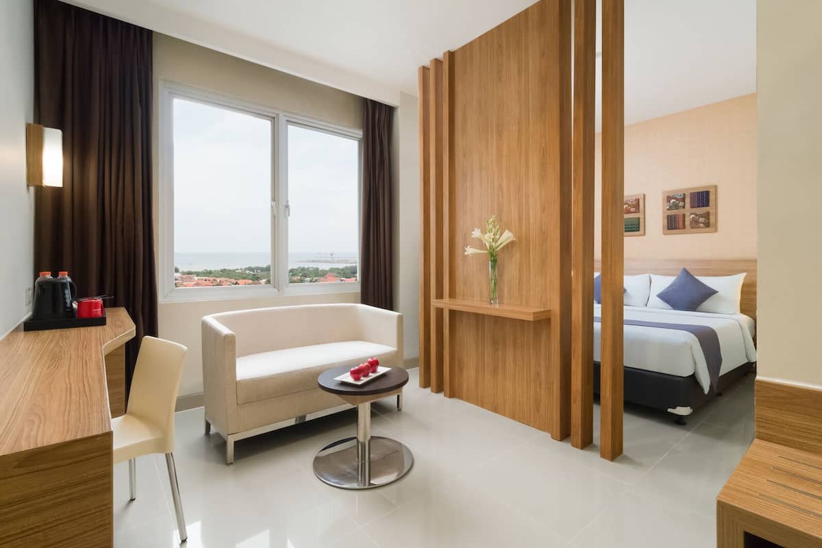 Une chambre d’hôtel moderne avec une grande fenêtre, un canapé, une petite table sur laquelle se trouve une assiette de pommes, un lit avec des oreillers et une cloison en bois.