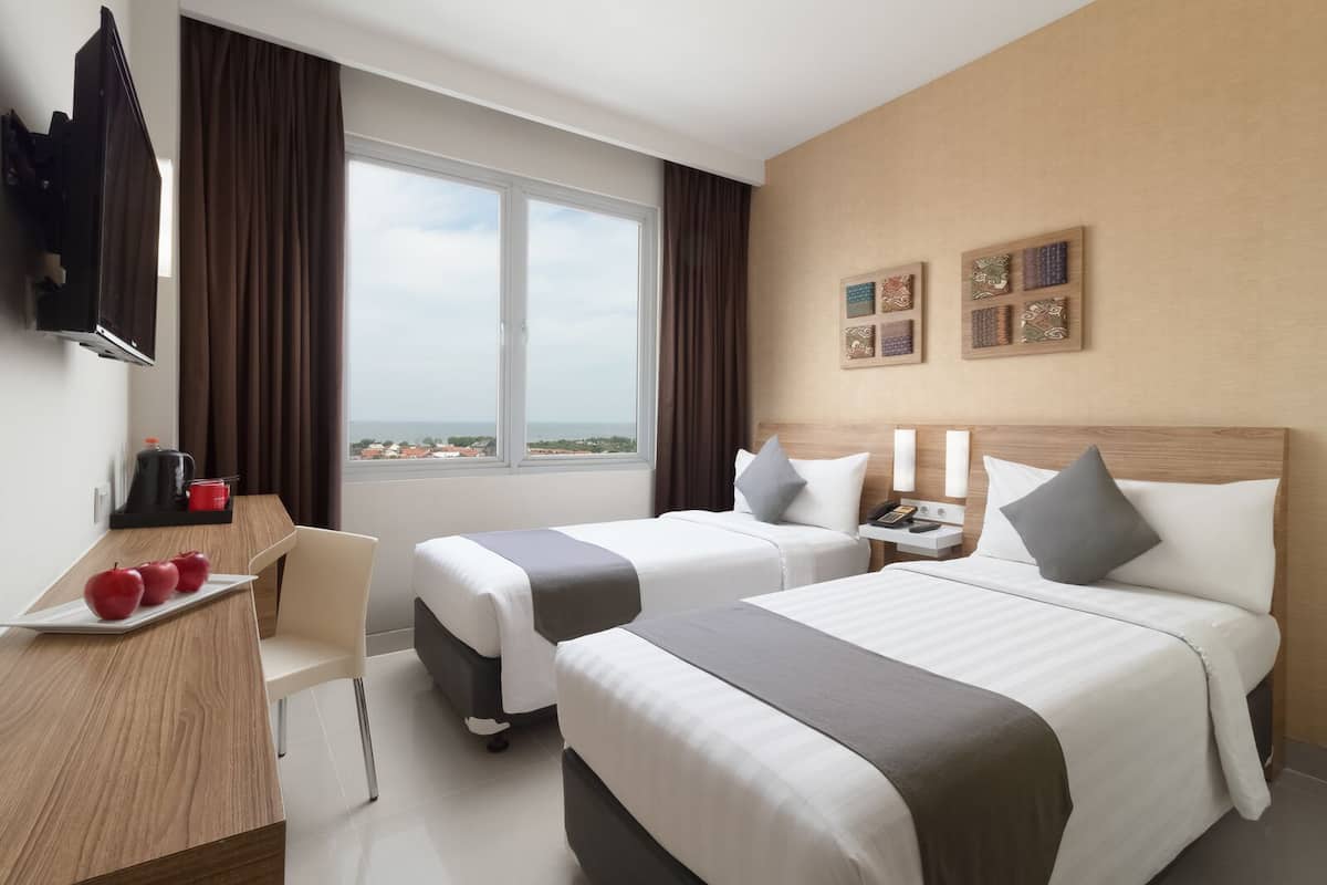 Une chambre d’hôtel avec deux lits, un bureau sur lequel se trouve une assiette de pommes, une télévision et une fenêtre donnant sur un paysage.