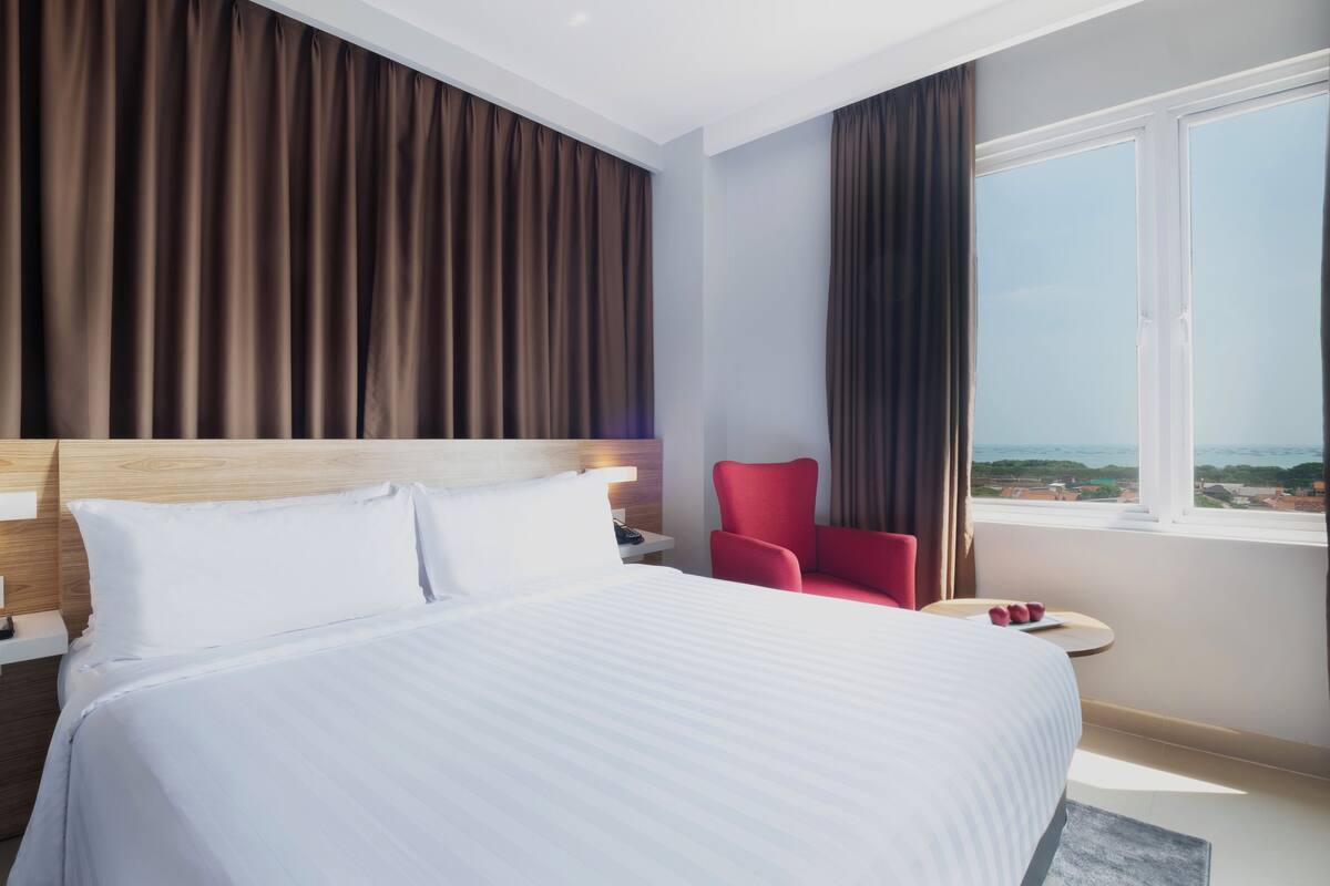 Une chambre d’hôtel avec un grand lit, un fauteuil rouge, une petite table et une fenêtre donnant sur un beau paysage.
