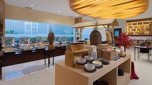 Un moderno área de desayuno tipo bufé con diversas estaciones de comida, un mostrador central de servicio y un espacio de estar con mesas y sillas.