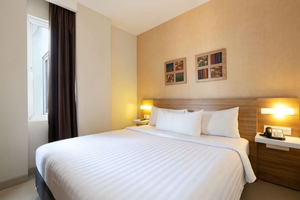 Habitación de hotel con una cama grande, dos mesitas de noche con lámparas, un cabecero y una ventana con cortinas.