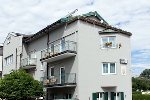 Un edificio de varios pisos con balcones, un letrero visible que indica un comercio y un árbol en la fachada.
