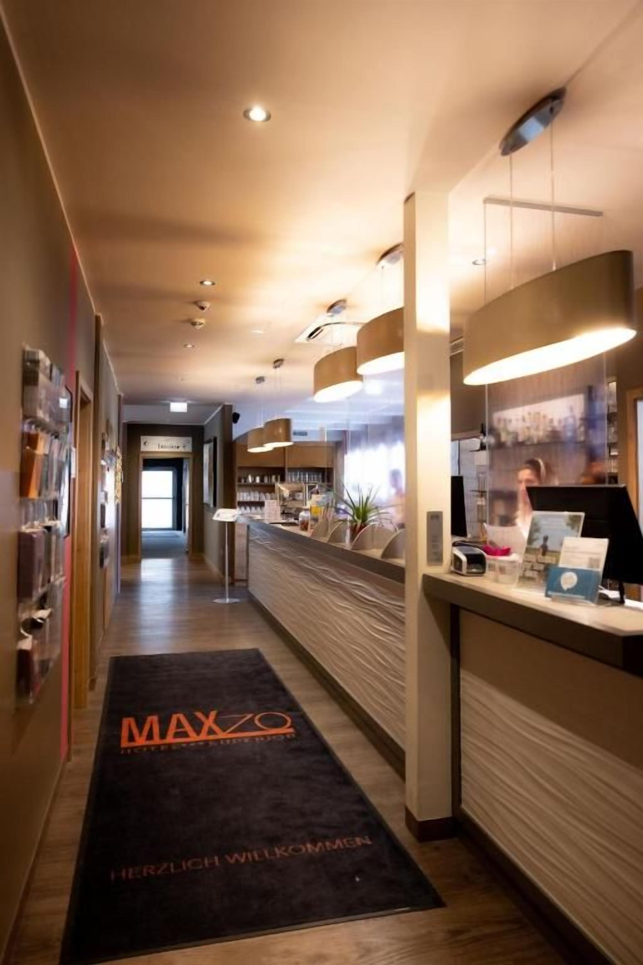Ein Hotel-Empfangsbereich mit einer Rezeption, einer schwarzen Willkommensmatte und einem Gang zum Eingang.