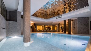 An indoor swimming pool with a modern design, featuring a clear pool area, a seating area with chairs, and a decorative ceiling.
