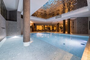 En indendørs swimmingpool med moderne design, med et klart bassin, et loungeområde med stole og et dekorativt loft.