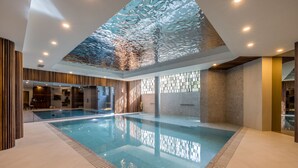 A modern indoor swimming pool with a glass ceiling, wooden paneling, and a clear water surface.