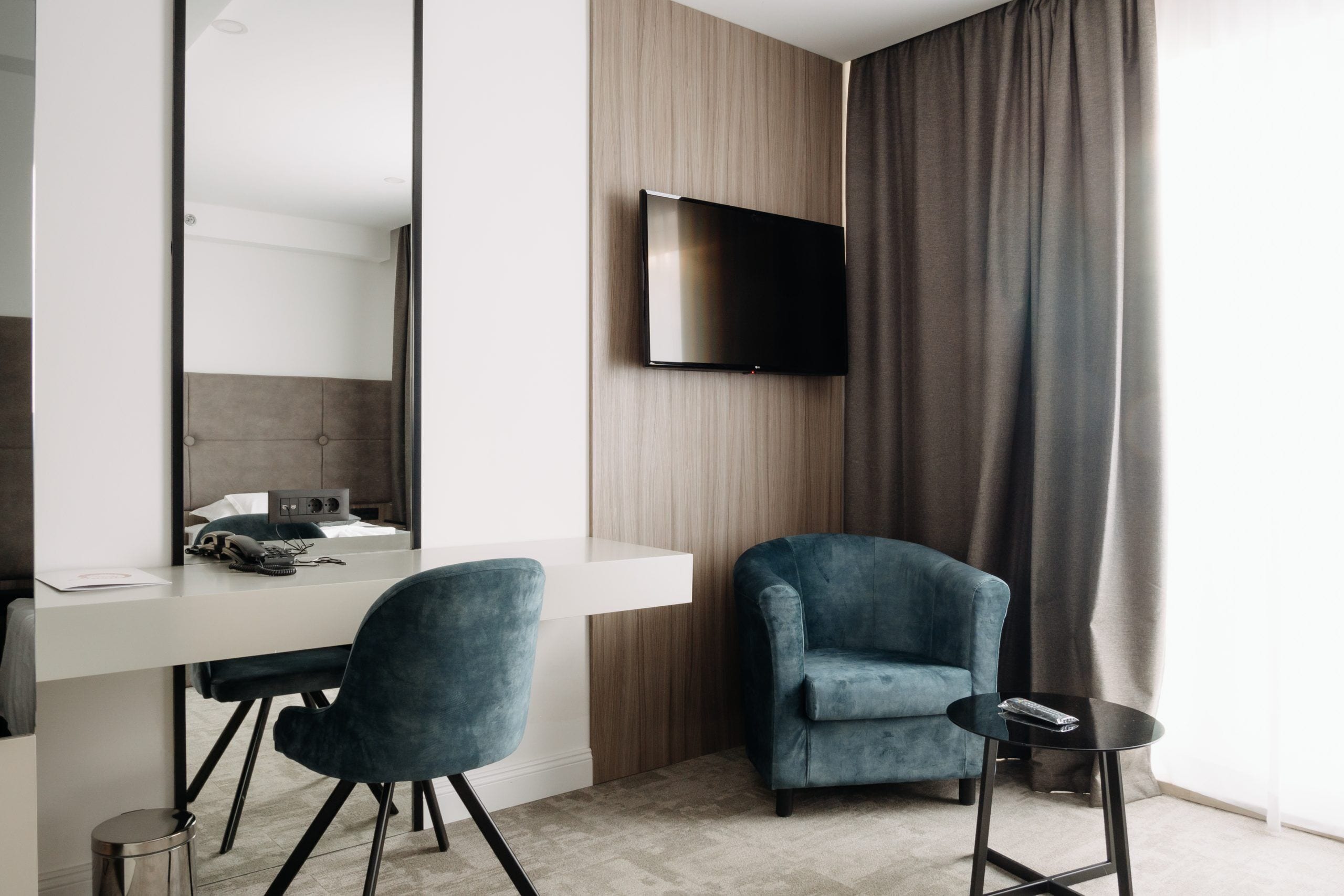 A modern hotel room with a flat-screen TV mounted on the wall, a desk with a chair, a small round table, and a blue armchair.