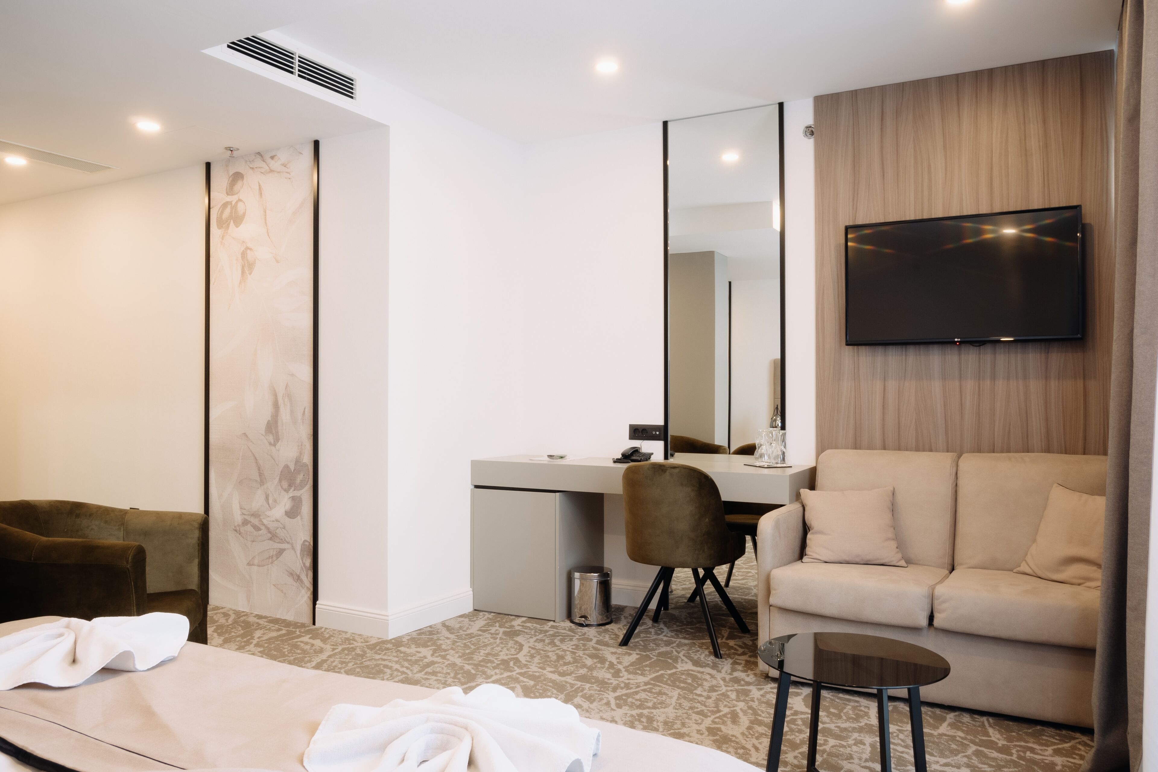 A modern hotel room with a bed, a desk, a chair, a sofa, a TV, and a small table.