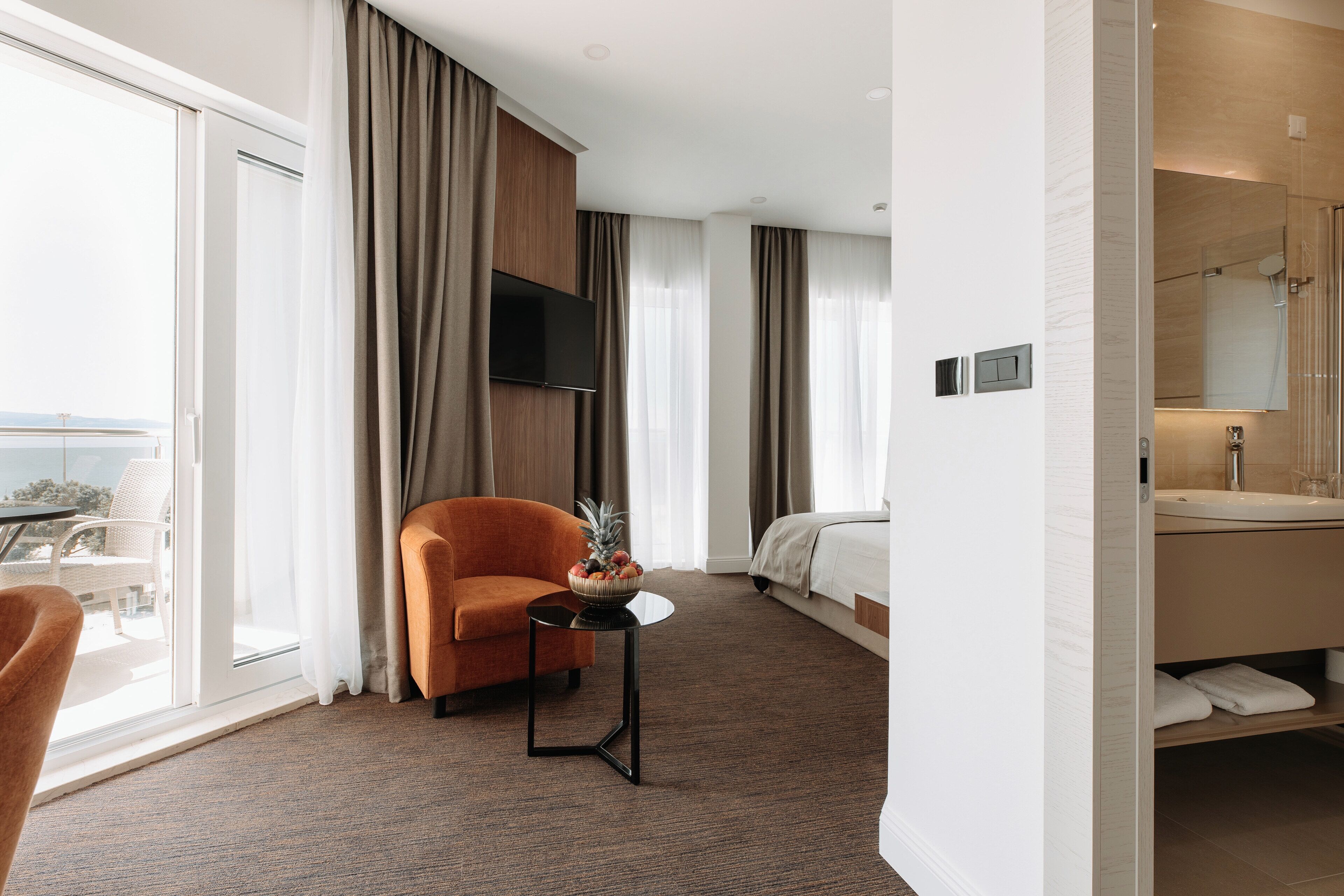 Une chambre d’hôtel comprenant un lit, un canapé, une petite table avec un panier de fruits, une salle de bain avec un lavabo et un miroir, et une vue sur l’extérieur grâce à une porte coulissante.