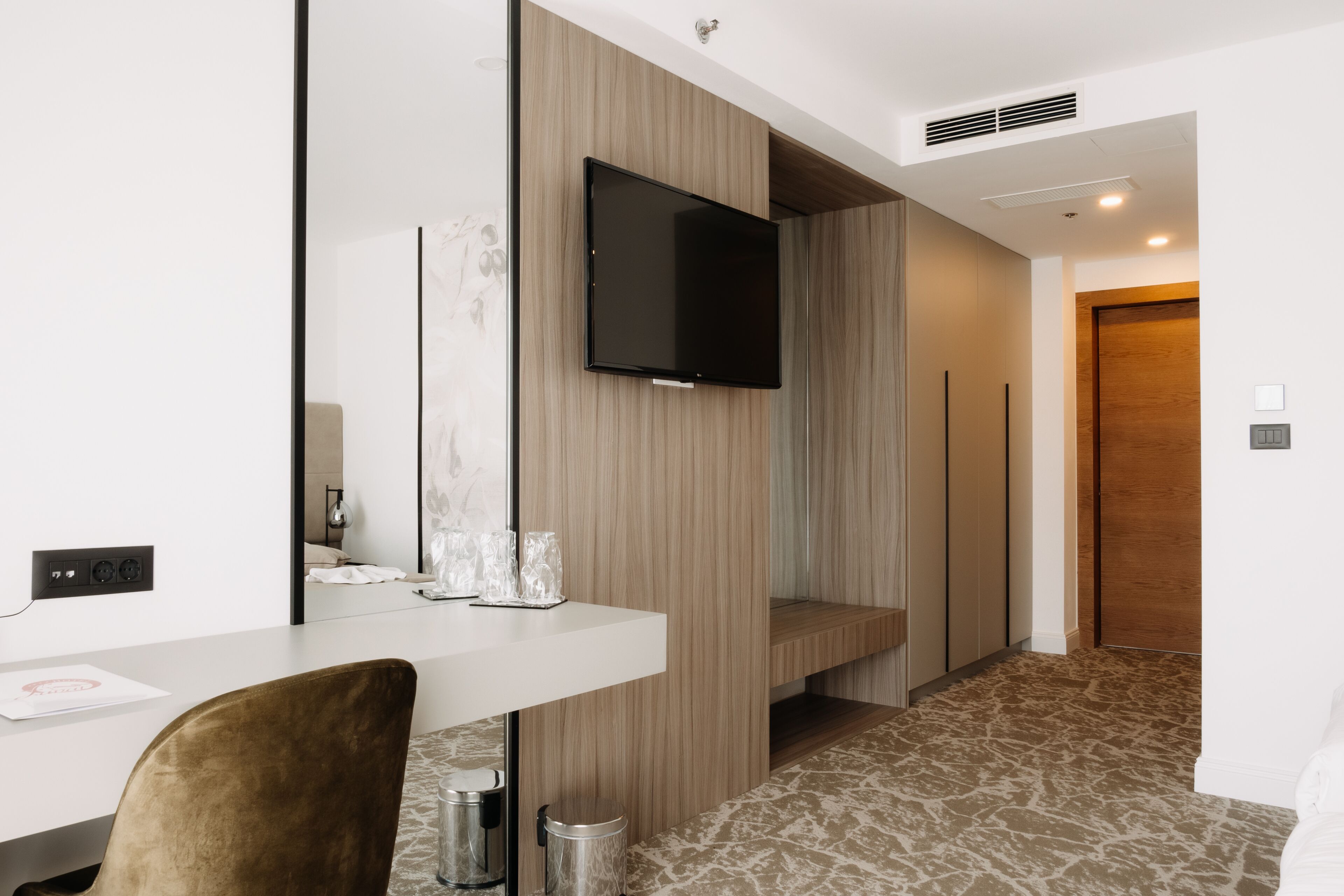 A modern hotel room with a flat-screen TV mounted on the wall, a wooden accent wall, a white desk with a chair, and a patterned carpet.