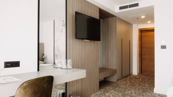 A modern hotel room with a flat-screen TV mounted on the wall, a wooden accent wall, a white desk with a chair, and a patterned carpet.