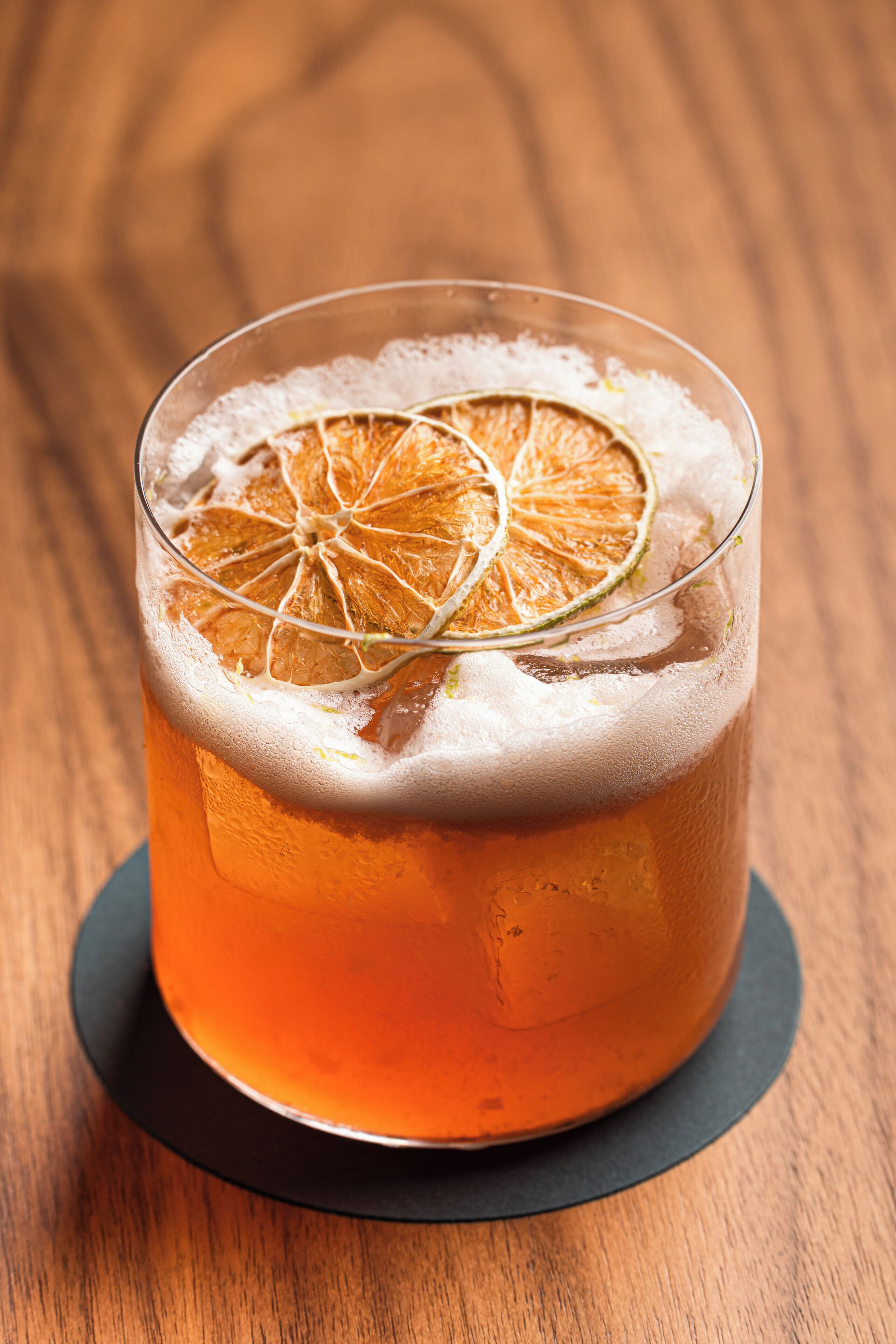 A glass of amber-colored beverage with a frothy head, garnished with a slice of citrus fruit, on a dark coaster.