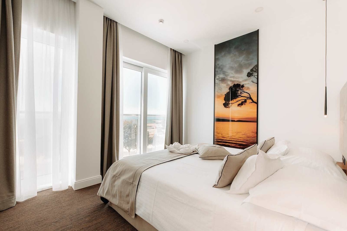 A hotel room with a large bed, a view of the sea through the window, and a painting of a tree on the wall.