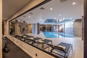 A modern indoor swimming pool with lounge chairs and a glass wall.