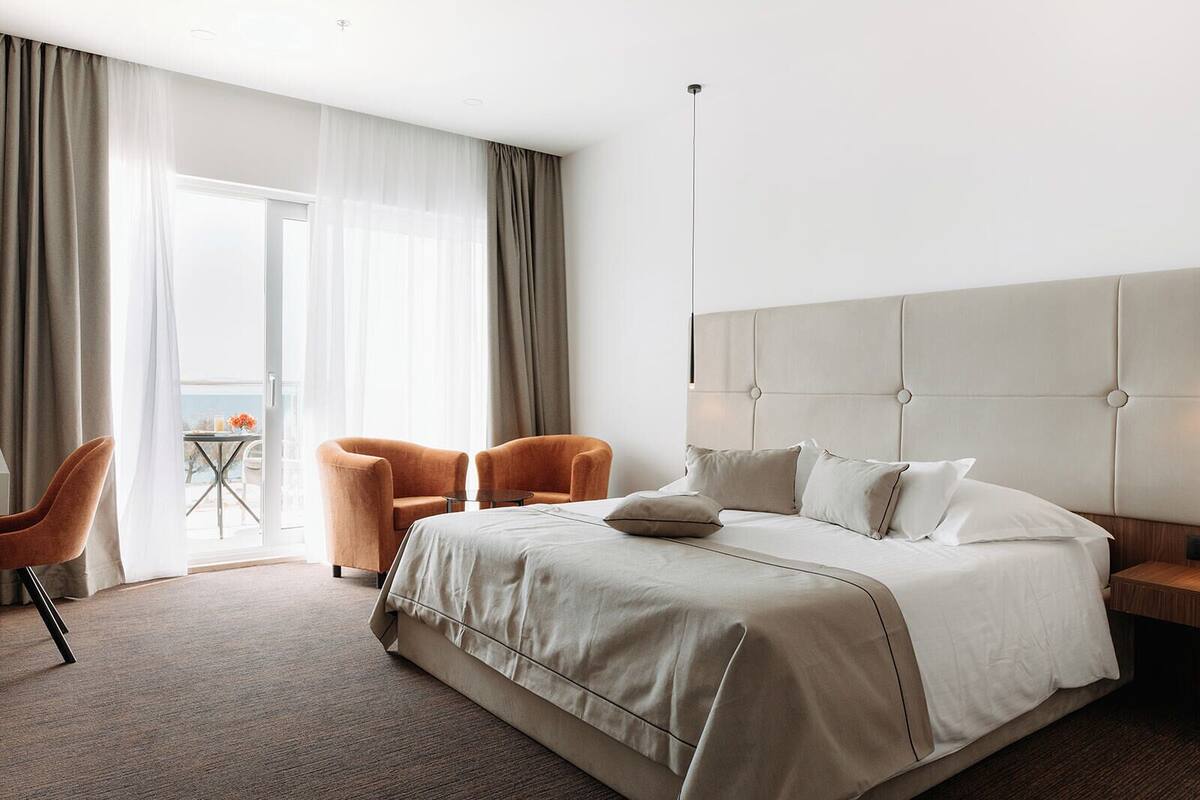 A modern hotel room with a large bed, two armchairs, a small table, and a balcony with curtains.