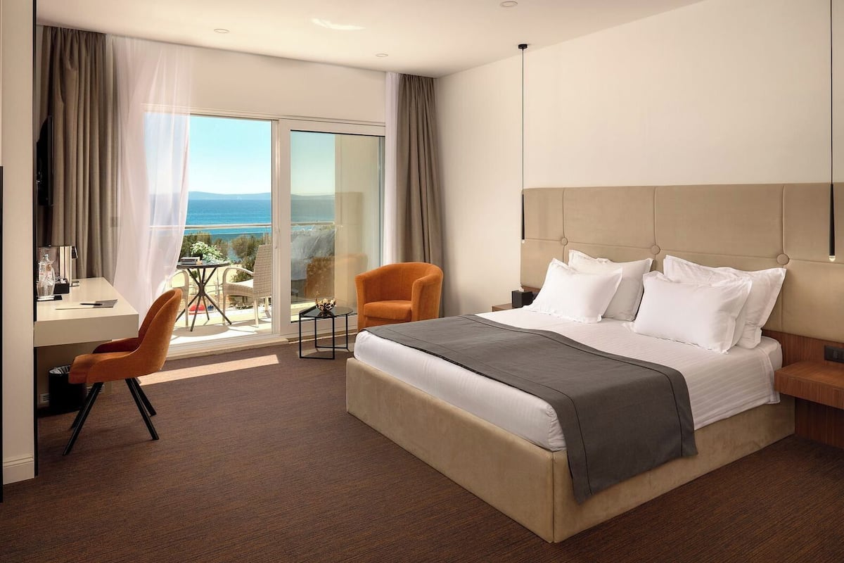 Ein modernes Hotelzimmer mit einem großen Bett, einem Schreibtisch mit Stuhl, einem kleinen Tisch und Blick auf den Ozean durch eine Glastür.