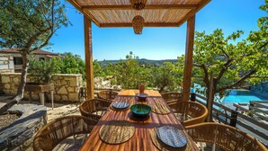 An outdoor dining area with a wooden table and wicker chairs, overlooking a swimming pool and a scenic view of hills. - Asion Lithos (Heraklion)