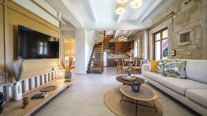 A spacious living area with a large flat-screen TV, a wooden bench, a round coffee table, and a sofa with patterned cushions. A staircase leads to an upper level with a kitchen and dining area. - Asion Lithos (Heraklion)