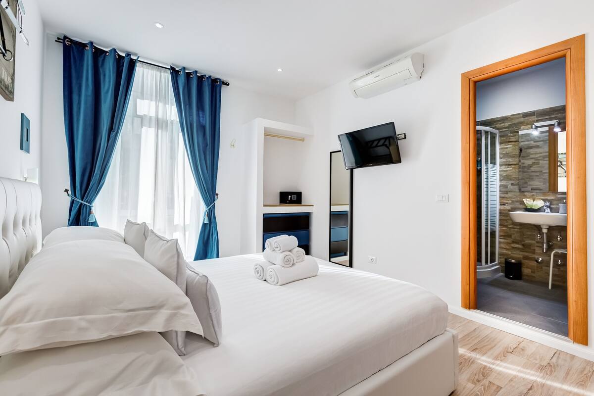 A hotel room with a bed, a TV, a bathroom visible through a glass door, and a wardrobe.