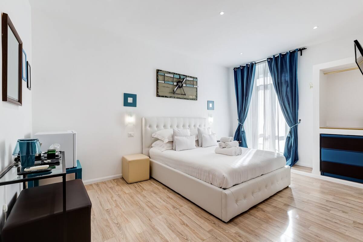 A modern bedroom with a large bed, a bedside table, a desk with a chair, a wardrobe, and a clock on the wall.