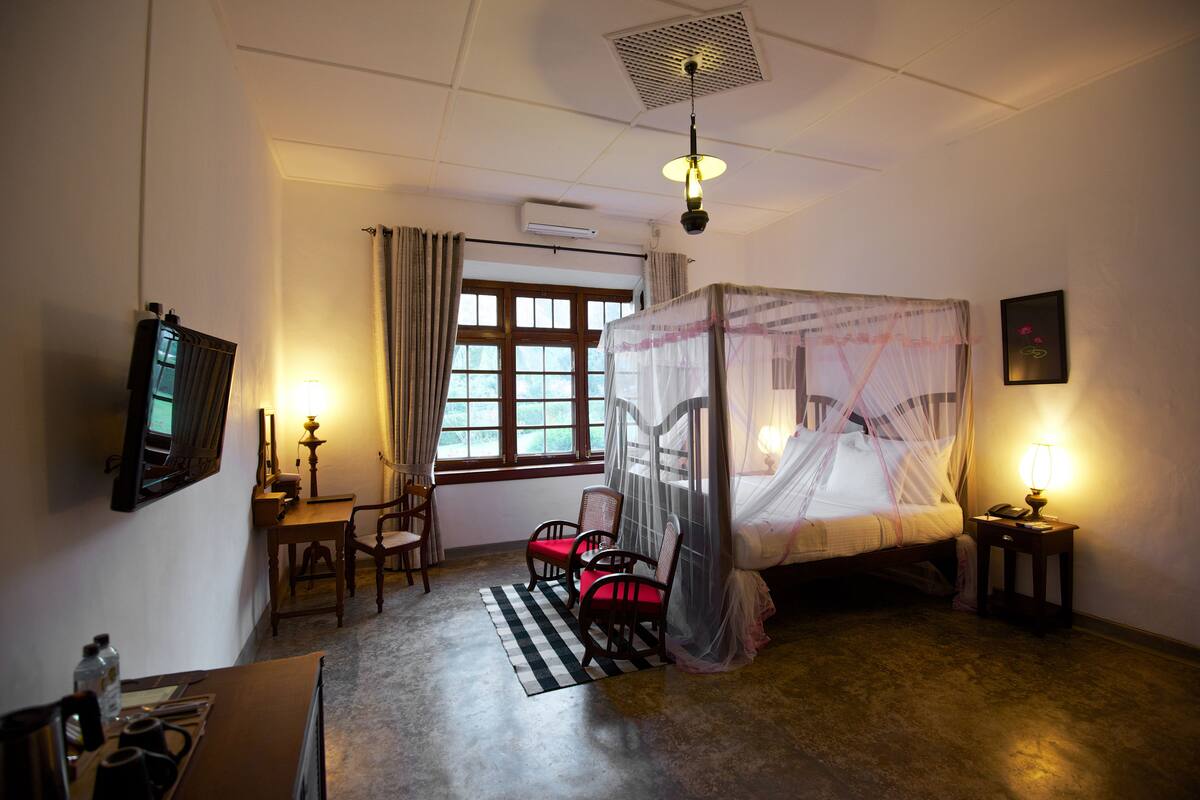 Un dormitorio con cama con dosel, un escritorio con silla, una mesita de noche con lámpara y una ventana con cortinas.