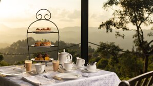 A table set for tea service with a three-tiered stand holding pastries, cups, and a teapot, overlooking a scenic landscape. - Mountbatten Bungalow (Kandy)