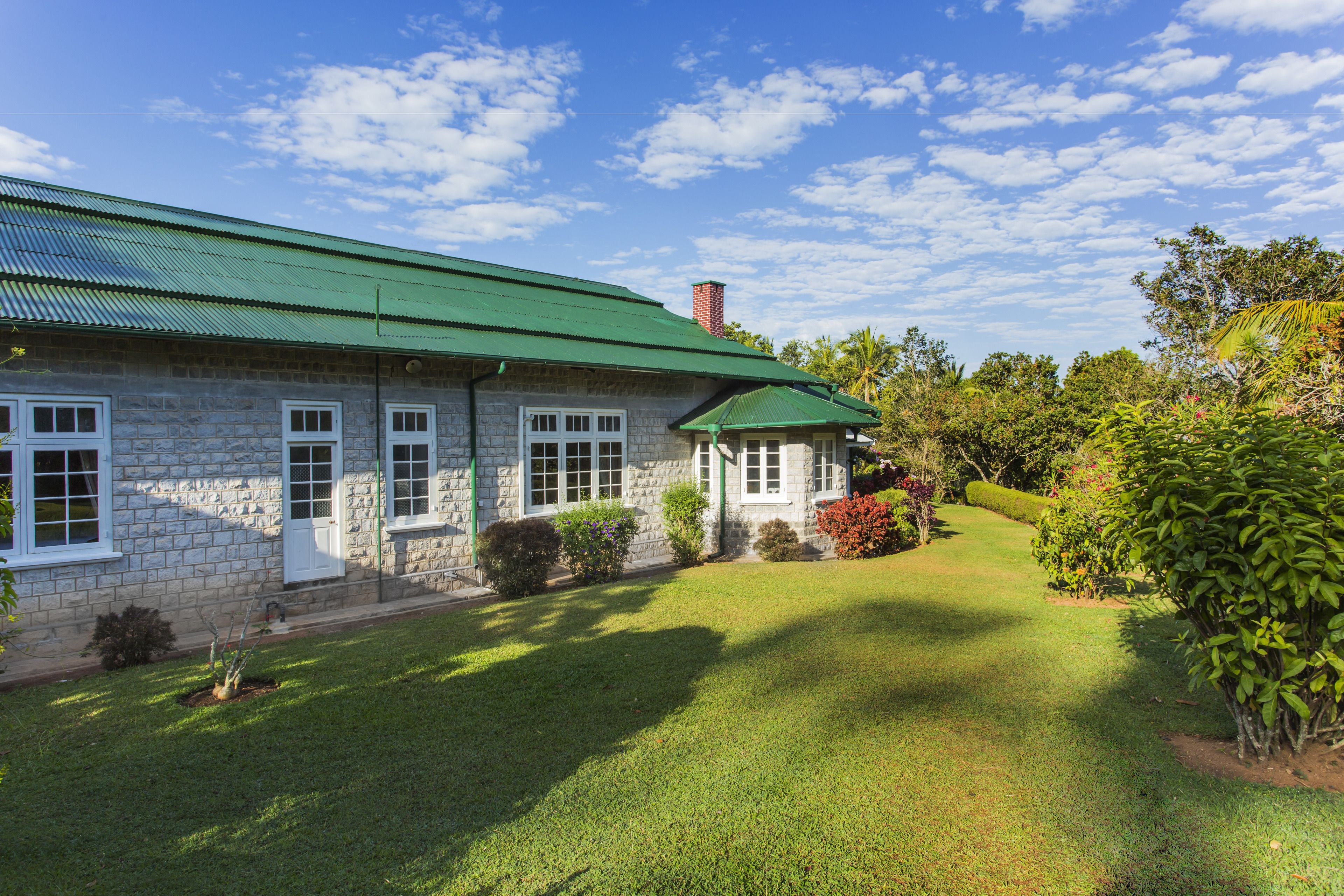 A stone house with a green roof and white windows, surrounded by a well-maintained lawn and various shrubs.