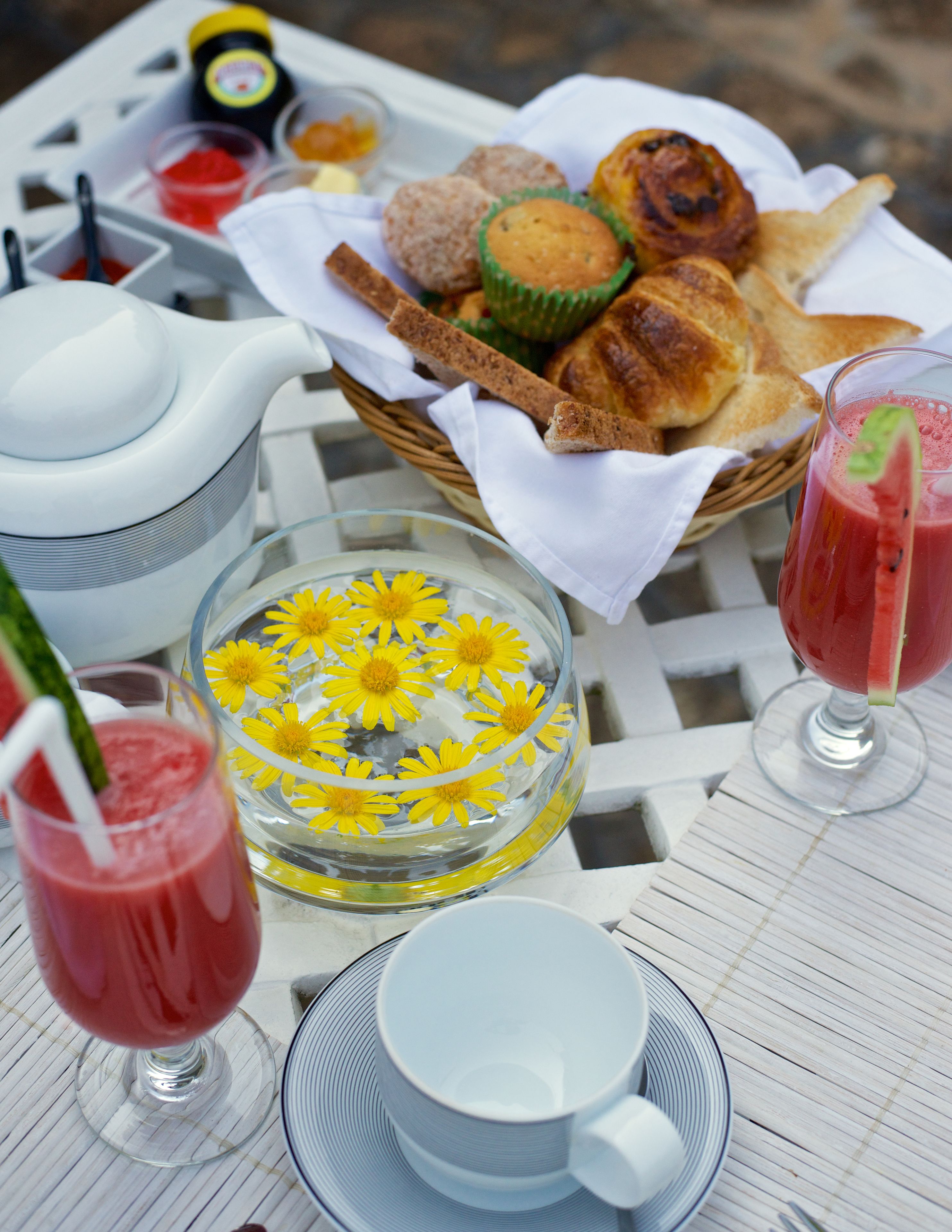 Una mesa de desayuno con una tetera, tazas, un vaso con flores, una cesta de pasteles y un vaso de bebida roja.