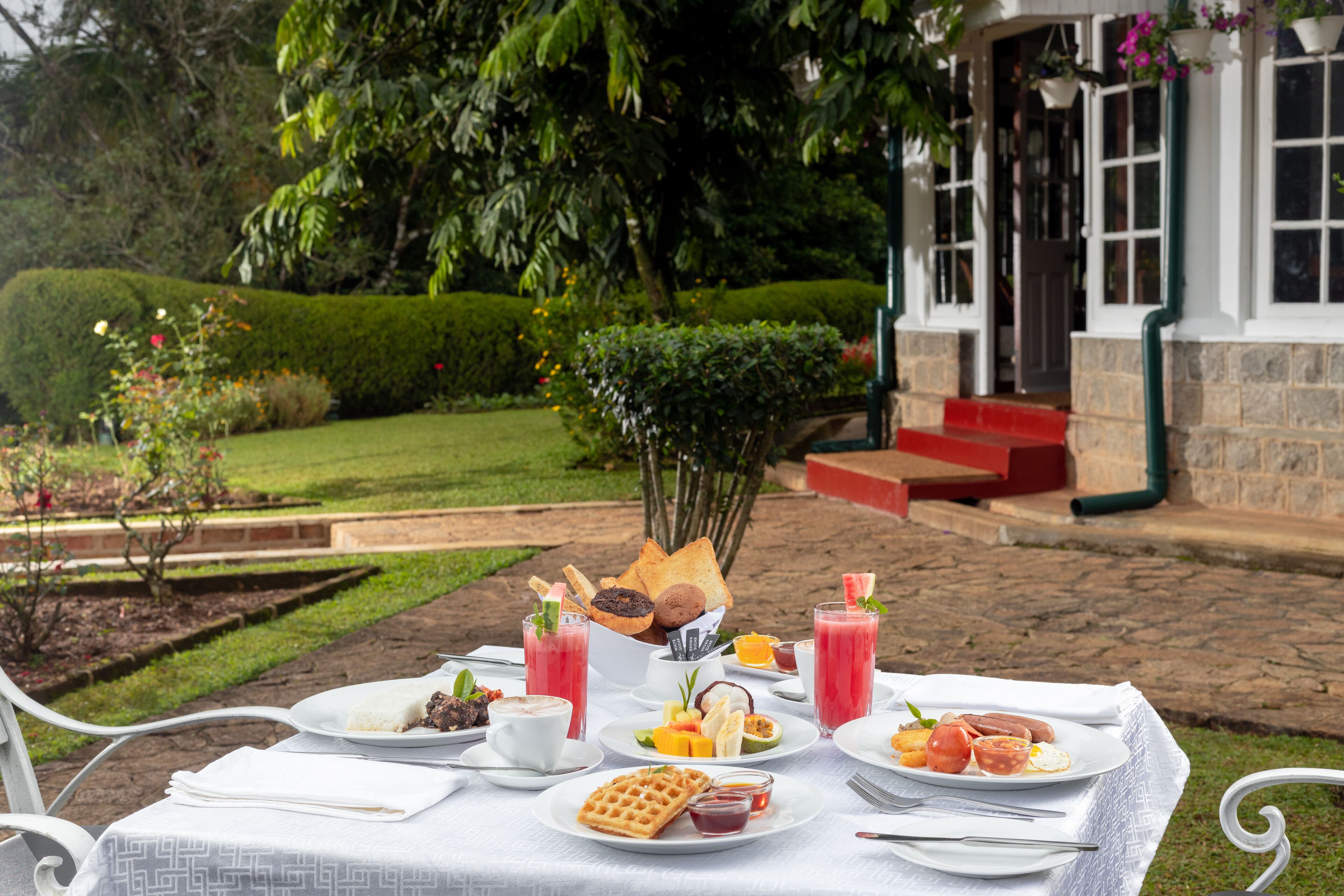 A table set outdoors with various breakfast items, including waffles, fruit, and beverages, in a garden setting with a building in the background.